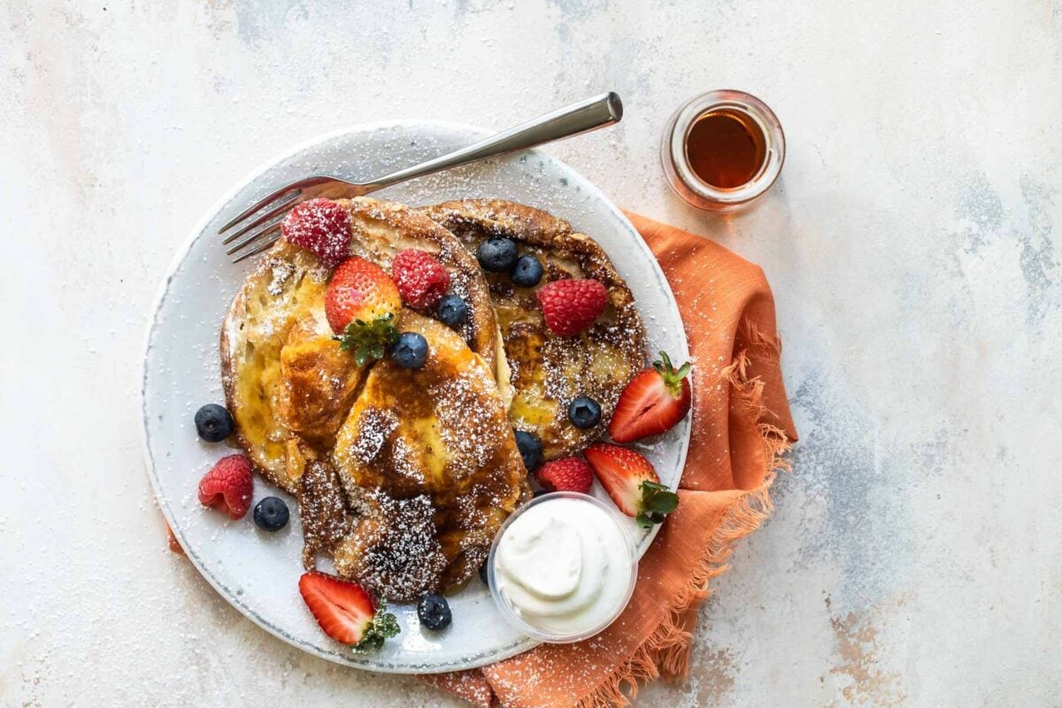 Croissant French toast on a white plate with fruit and whipped cream.