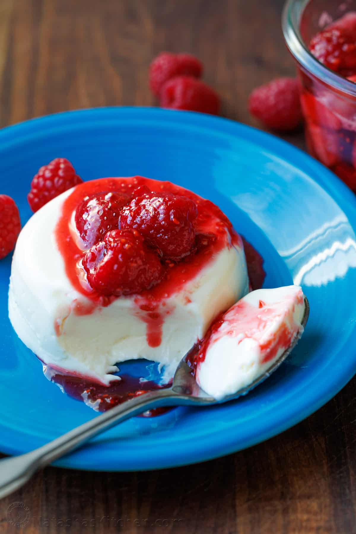 Panna Cotta with fresh berry sauce cascading down the side with one bite loaded onto a spoon laying on a blue dessert plate.